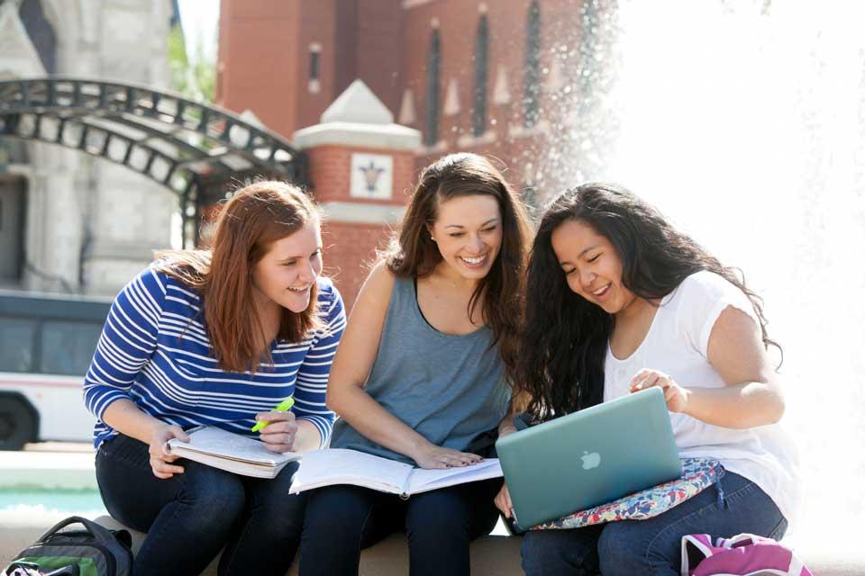 Three students share a look at a laptop, with notebooks also on their laps, while sitting in front of a fountain.